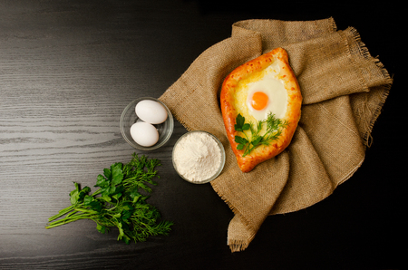 Khachapuri with eggs on sackcloth, flour, eggs and parsley on black table, space for textの写真素材