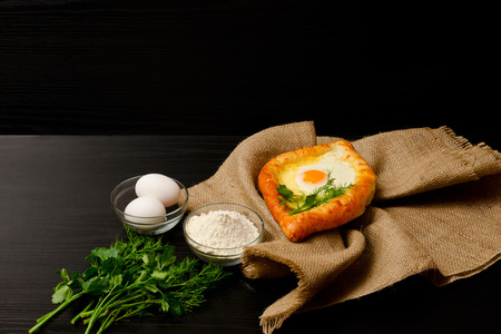 Khachapuri with eggs on sackcloth, flour, eggs and parsley on black table, space for textの写真素材