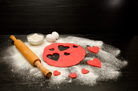 Red dough, cut out hearts, flour, eggs and rolling pin on a black background, space for textの写真素材
