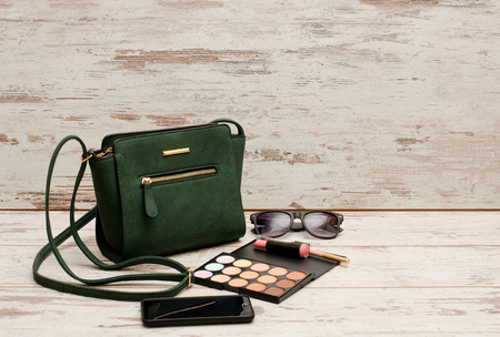 Green ladies handbag, sunglasses, phone, eye shadow and lipstick on wooden background. Fashion concept with space for textの写真素材
