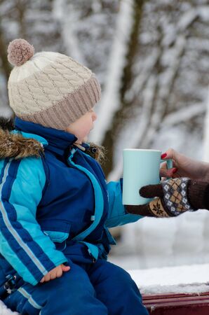 Boy in winter clothes taking a mug of female hands on a background of snowy treesの写真素材