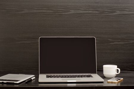 Empty laptop screen, graphic tablet, phone and mug on a black wooden background