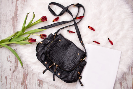 Black bag, a white sheet of paper and withered tulips on white fur. Fashionable concept, top view