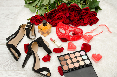 Red carnival mask, perfume, shoes and lipstick on a wooden table on a background of scarlet rosesの写真素材