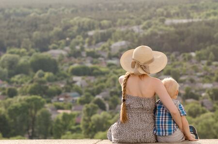 Mom and son sitting on the background of green forest and the villageの写真素材