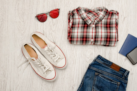 Red and white checkered shirt, glasses, sneakers, jeans, phone and passport. Wooden background. Fashionable concept, top viewの写真素材