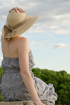 Beautiful girl in a hat and dress sits on a background of a cloudy blue sky and forestの写真素材