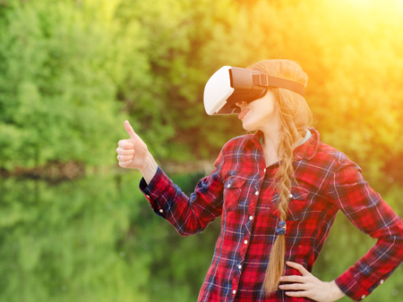 Girl shoots virtual reality glasses, sunset in nature, back view. Thumbs up