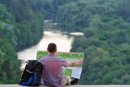 Man with backpack and open topographic map sits on the background of river and green forestの写真素材