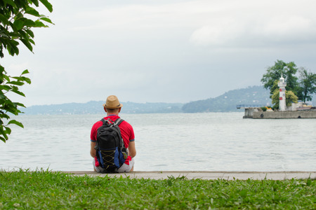 Young man in a hat and with a backpack is sitting on the pier on the background of the sea, lighthouse and mountains in the distance. View from the backの写真素材