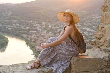Beautiful girl with backpack in a wide hat sitting on background of the river, mountains and the city belowの写真素材