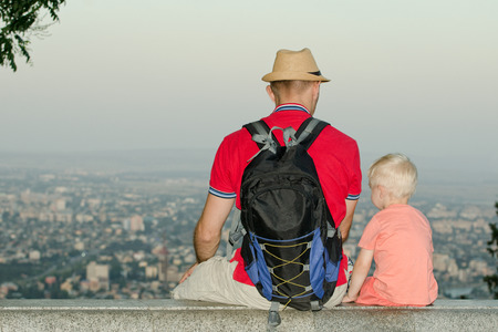 Young man in backpack and hat sitting with son on a background of a big city at the bottom. Back viewの写真素材