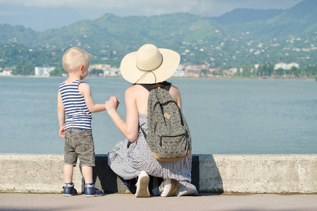 Mom and her son at the pier on the sea background, lighthouse and mountains in the distance. Back viewの写真素材