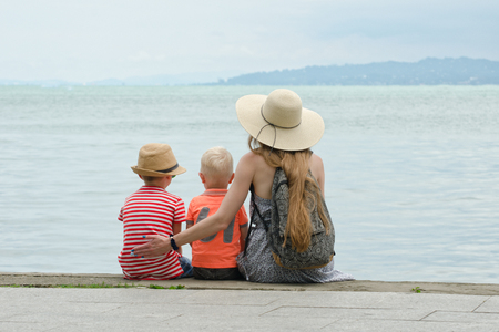 Mom and two sons sit on the pier and admire the sea and the mountains in the distance. Back viewの写真素材