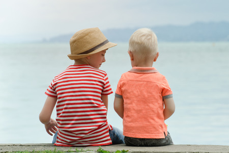 Two little brothers sit on a pier against the sea and mountains in the distance. Back viewの写真素材