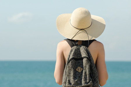 Girl in a hat with a backpack standing on the coastline. View from the backの写真素材