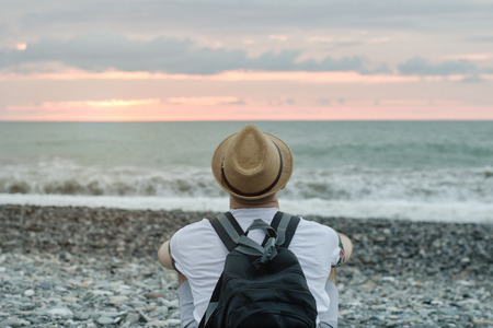 Young man in hat and with backpack sitting on the beach against the backdrop of the sea and sunset sky. Back viewの写真素材
