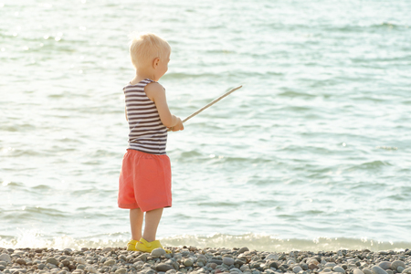 Little boy in a striped T-shirt is standing on the seashore with a wand in his hands. View from the backの写真素材