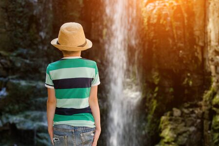 Boy in a hat looking at a waterfall. View from the backの写真素材
