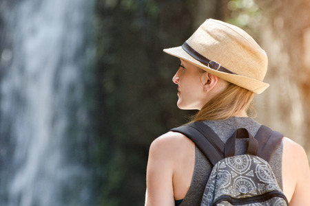 Girl in a hat with backpack looking at a waterfall. View from the backの写真素材