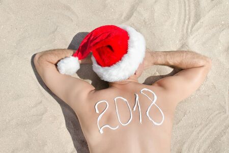 Man in Santa hats with the inscription 2018 on the back lying on the sand. View from above