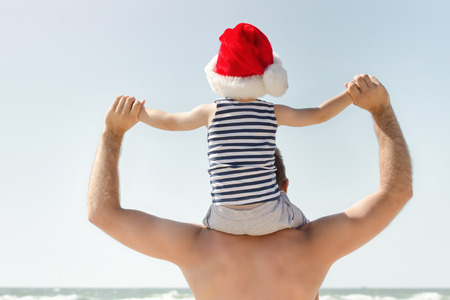 Kid in Santa hat sitting on his father's shoulders. Sea shore. Back viewの写真素材