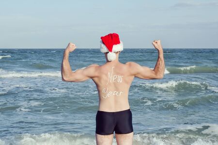 Strong man in Santa hats with the inscription New Year on the back standing on the beach. Back viewの写真素材