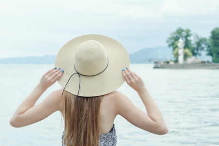 Girl in a hat on the pier. Mountains and lighthouse on the background. View from the back