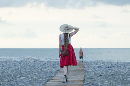Girl in red skirt and hat walking along the deck to the sea, sunset. View from backの写真素材