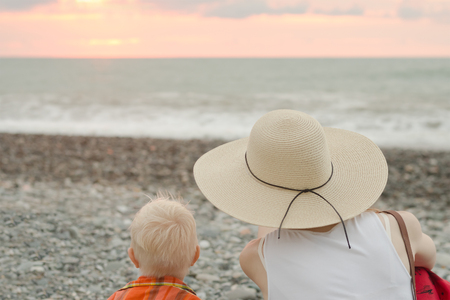 Mom and son rest on the pebble beach. Sunset time. Back viewの写真素材