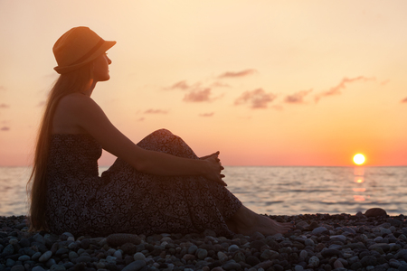 Girl in a hat sitting on a background of the sea at sunset. Silhouetteの写真素材