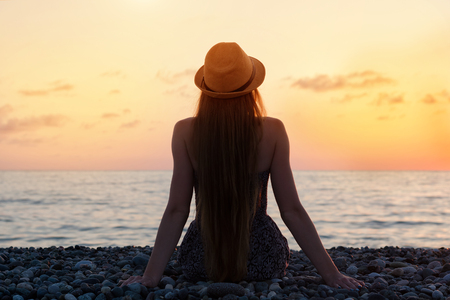 Girl in the hat sitting on the seashore. Sunset time. Silhouette.  Back viewの写真素材