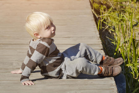 Blond little boy is sitting on the pier. Autumn sunny day. River bankの写真素材