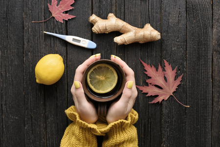 Mug of tea in female hands. Lemon, ginger and thermometer. Cold treatment. Wooden background. Top viewの写真素材