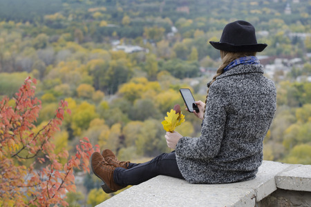 Girl with a phone sits on a hill, yellow leaves in her hand. Autumn on the backgroundの写真素材