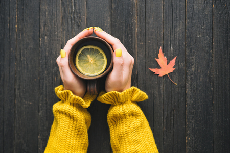 Mug of tea with lemon in a female hand. Wooden background, autumn leaf. Top viewの写真素材