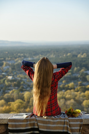 Young woman with long hair sits on a hill overlooking the city. Back viewの写真素材