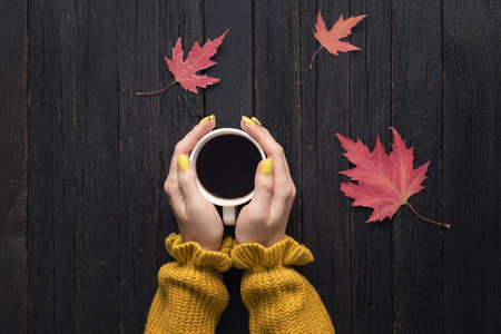 Mug of coffee in a female hand. Wooden background, autumn leaves. Top viewの写真素材