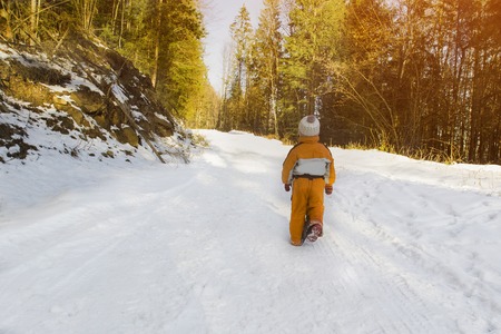 Little boy in an orange jumpsuit walking on snow-covered road in a coniferous forest. Winter sunny day. Back view.の写真素材