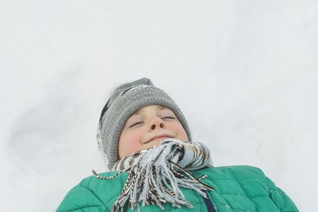 boy in a a hat, scarf and a green jacket is lying on his back in the snow with eyes closed. Portrait. Close-up. Winter dayの写真素材
