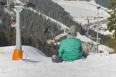 Boy sits on a snow-covered hill on background of cable car, pine forest and mountains. Ski resort. Winter day.の写真素材