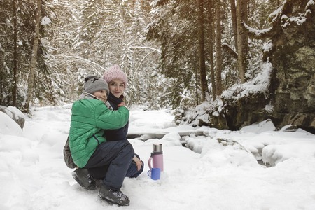 Mom and son are sitting and hugging against the backdrop of snow-covered forest. Smiling faces. Winter cloudy dayの写真素材