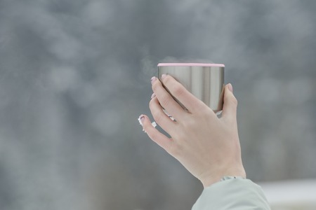 Glass from a flusk with steaming drink in a female hand on a blurred background. Snowflakes on the fingers.の写真素材