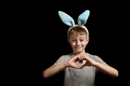 Blond boy in hare ears folded his hands in heart shape on his chest on black background. Love and family concept. Copy space.の写真素材