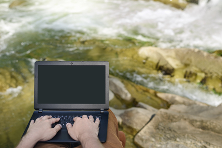 Work in nature concept. Men's hands on the laptop keyboard on the background of a mountain river. Place for text on the screenの写真素材