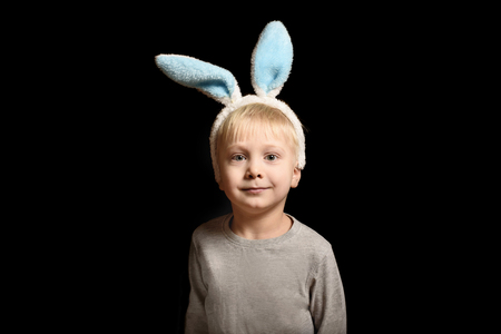 Cute little blond boy in the hare's ears standing on a black background. Portrait. Copy space.