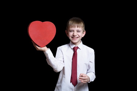 Laughing fair-haired boy in a shirt and tie holding a red heart shaped box. Love and family concept. Isolate on black backgroundの写真素材