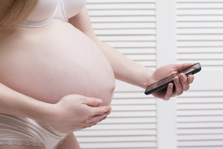 Pregnant woman in underwear holding hand on stomach and uses smartphone. Call an ambulance. Prenatal contractions.の写真素材