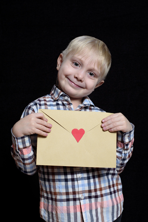 Smiling little blond boy holds an envelope with a heart. Isolate on black backgroundの写真素材