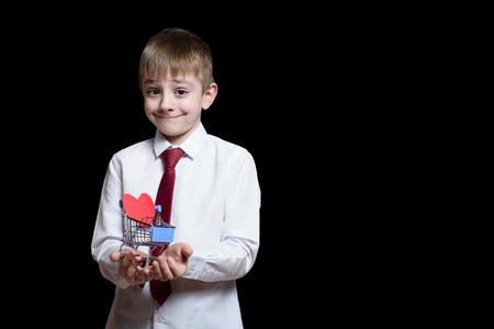 Smiling boy in light shirt and tie holds a metal shopping trolley with a heart-shaped postcard inside. Isolate on black backgroundの写真素材